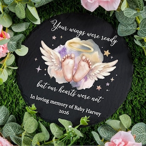 May include: A round black slate memorial stone with the text "Your wings were ready but our hearts were not". It features baby feet with angel wings, a halo, and stars. The text "In loving memory of Baby Harvey 2026" is also included.