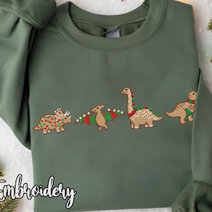 Może przedstawiać: Oliwkowo-zielona bluza z haftowanym wzorem czterech piernikowych dinozaurów. Dinozaury zdobione są czerwonymi, zielonymi i białymi akcentami. Słowo "Embroidery" jest napisane kursywą na dole.