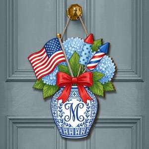 May include: A patriotic door hanger featuring an American flag, blue hydrangeas, and red and white fireworks. The design includes a red bow and a blue and white vase with the letter "M". The door decoration is hanging from a gold door knocker.