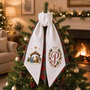 May include: A white fabric tree topper featuring two long, pointed tails. One tail displays an embroidered nativity scene with a star, while the other features a red embroidered letter P surrounded by a green and red holly leaf wreath.