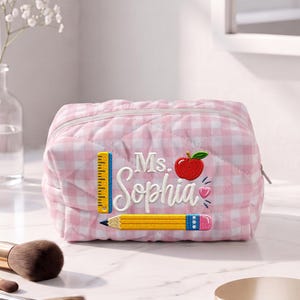 May include: A quilted pink and white gingham zippered pouch featuring embroidered designs of a yellow ruler, a red apple, a yellow pencil, and a small pink heart. The text "Ms. Sophia" is embroidered in white script in the center.
