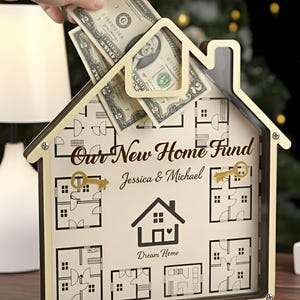 Op de afbeelding: Een houten huisvormige spaarpot met een doorzichtige voorkant. De tekst "Our New Home Fund" en de namen "Jessica & Michael" staan erop. Een hand steekt bankbiljetten in de gleuf aan de bovenkant.