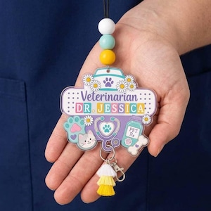 Puede incluir: Un portatarjetas con un dije de acrílico en forma de venda con el texto Veterinarian DR. JESSICA, decorado con margaritas, un estetoscopio, una cara de gato, una huella y un frasco de medicina, acentuado por una borla blanca, amarilla y naranja.