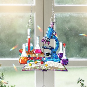 Puede incluir: Una ventana colgante de vitral con un microscopio, vasos de precipitados y un libro. El microscopio es azul y plateado, los vasos de precipitados están llenos de líquidos de colores y el libro tiene un diseño de arcoíris.