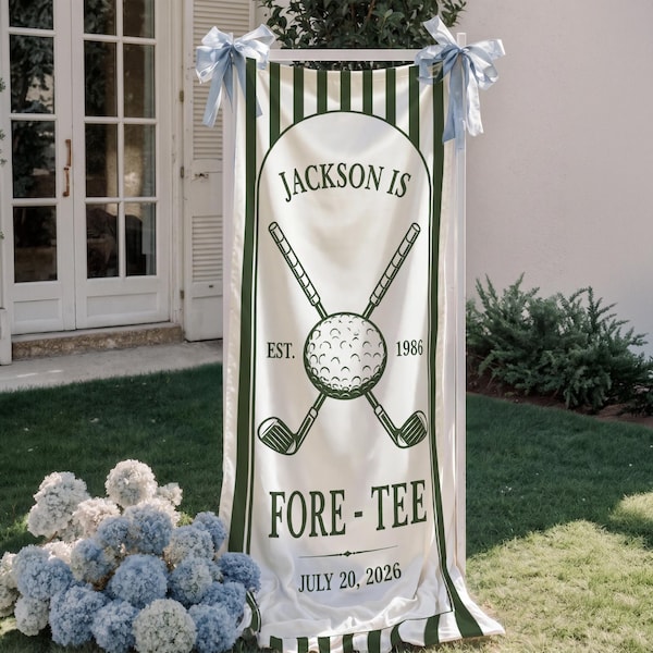 May include: A white banner with green stripes and text that reads "JACKSON IS FORE-TEE JULY 20, 2026." The banner features a golf ball and crossed golf clubs. Light blue ribbons are tied at the top.
