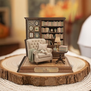 May include: A miniature diorama featuring a beige tufted armchair with a green throw blanket, a wooden bookshelf filled with books, a small side table with a lamp and teacup, and a plaque reading JUST A GIRL WHO LOVES BOOKS EMMA.
