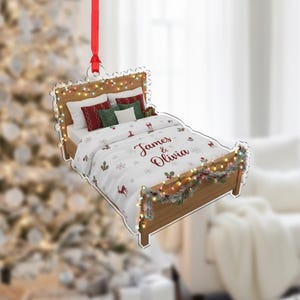 May include: A Christmas ornament shaped like a bed, with a wooden frame and a white comforter. The comforter has a pattern of snowflakes, deer, and holly. The names "James & Olivia" are written on the comforter. The bed is decorated with string lights and a red ribbon.