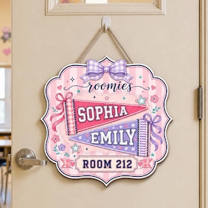 May include: A decorative wall sign with a light pink striped background, featuring a purple bow at the top. It displays the text roomies, the names SOPHIA and EMILY on two pennants, and ROOM 212 at the bottom, decorated with small stars and flowers.