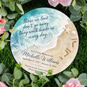 Pode incluir: Uma placa memorial redonda com uma cena de praia em aguarela com ondas azuis, areia, uma estrela-do-mar e uma bolacha-da-praia. O texto diz: Those we love don't go away, they walk beside us every day. In Loving Memory of Michelle Williams.
