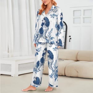 Puede incluir: Conjunto de pijama blanco con un estampado de perro y flores azules. La parte superior de manga larga tiene una botonadura y cuello con solapas. Los pantalones son de corte recto. La tela parece ser un material suave y sedoso.