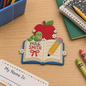May include: An embroidered patch featuring an open book with the text "mrs. SMITH" in black and red, a red apple, a green caterpillar wearing glasses, a yellow pencil, and small white daisy flowers with yellow centers on a blue border.