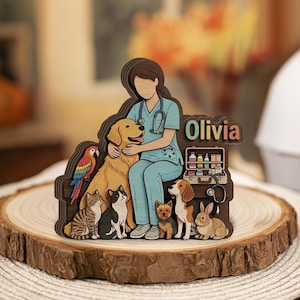 Peut inclure: Une découpe en bois multicouche représentant un vétérinaire en tenue bleu clair avec un stéthoscope, entouré d'un golden retriever, d'un perroquet, de deux chats, d'un petit chien, d'un beagle et d'un lapin. Le prénom Olivia est inscrit en lettres colorées.