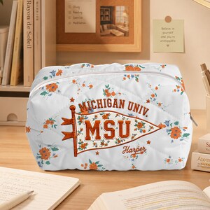 May include: A white quilted pouch featuring an all-over floral print with orange and blue flowers. Centered on the front is a graphic of a pennant flag with the text MICHIGAN UNIV. and MSU in orange, with the name Harper printed below it.
