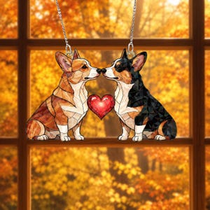 May include: A hanging ornament featuring two Corgi dogs with a geometric stained-glass style pattern. One dog is tan and white, the other is black, tan, and white. They face each other with a red heart shape positioned between their noses.