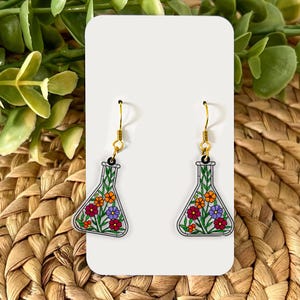 May include: A pair of earrings featuring a floral design. Each earring is shaped like a laboratory flask, filled with colorful flowers in shades of red, orange, and purple. The earrings have gold-colored hooks.