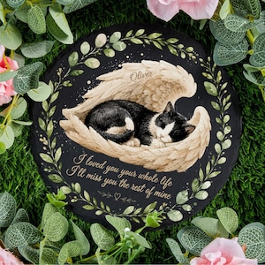 May include: A circular black memorial stone with a sleeping black and white cat in angel wings. The name "Oliver" is above the cat. The stone is framed by a green leaf border and the inscription reads: "I loved you your whole life, I'll miss you the rest of mine."