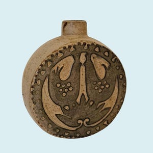 May include: A round, ceramic vase with a short neck. The vase is primarily tan with a circular, raised design in shades of green and tan. The design features stylized shapes and patterns, including a central figure and surrounding motifs. The vase has a rustic, handcrafted appearance.