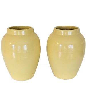May include: Two matching, light yellow ceramic vases. Each vase has a rounded body and a slightly flared rim. The vases have a glossy finish and are set against a white background. The overall shape is reminiscent of a classic urn.