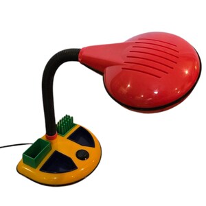May include: A retro-style desk lamp with a red, rounded shade and a flexible black neck. The base is yellow with green and blue accents, and includes a small storage compartment. The lamp is designed for desk use.