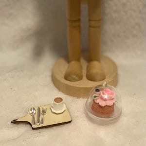 May include: A wooden nutcracker figurine with a hat and a round base. A miniature wooden serving board with a cup of coffee and silverware, and a cupcake under a clear dome are in the foreground.