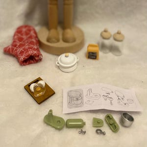 Könnte beinhalten: Eine Holz-Nussknacker-Figur mit hohem Hut und detailliertem Design. Das Bild enthält auch Miniatur-Küchenzubehör wie einen roten Ofenhandschuh, einen kleinen Topf und ein Schneidebrett mit Spiegelei.