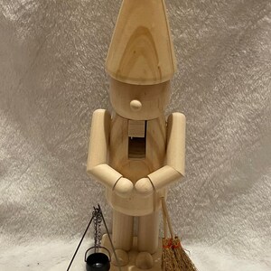 Puede incluir: Figura de madera de un gnomo con un sombrero cónico, con las manos juntas. Tiene un pequeño caldero negro, una escoba y tres pequeñas calabazas naranjas. La figura es de madera clara.