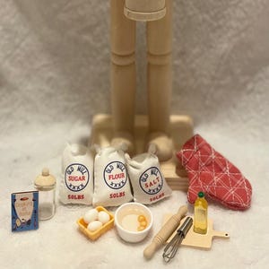 Könnte beinhalten: Ein hölzerner Nussknacker mit Trommelstöcken, umgeben von Miniatur-Backutensilien. Enthält Beutel mit Zucker, Mehl und Salz sowie Eier, eine Schüssel und einen roten Ofenhandschuh. Eine charmante, handgefertigte Küchen-Themenausstellung.