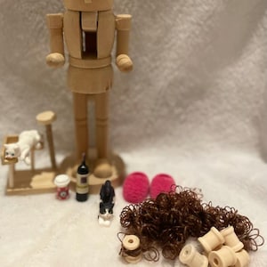 Op de afbeelding: Een houten notenkrakerpop met een hoge hoed, naast miniatuuraccessoires. De accessoires omvatten een kat op een kattenboom, een fles wijn, een koffiekopje, een zwart-wit kattenbeeldje, roze slippers, bruin haar en houten spoelen.