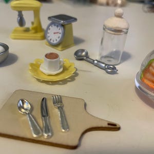 Könnte beinhalten: Miniatur-Küchenszene mit einem gelben Mixer, einer Waage und einer kleinen Tasse Kaffee auf einem gelben Teller. Enthält ein Schneidebrett mit Utensilien, ein Glas und ein Dessert unter einer transparenten Kuppel. Eine Schüssel mit Eiern ist ebenfalls vorhanden.