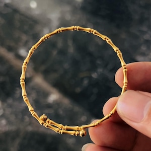 May include: A gold-coloured bracelet with a bamboo-style design. The bracelet is made of multiple connected segments, creating a flexible, circular shape. The bracelet is held by a hand against a dark, blurred background.