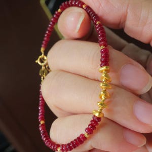 May include: A delicate bracelet featuring small, round, deep red beads interspersed with gold-colored accents. The bracelet has a gold clasp and a section of larger, irregular gold beads. The jewelry is held in a hand.