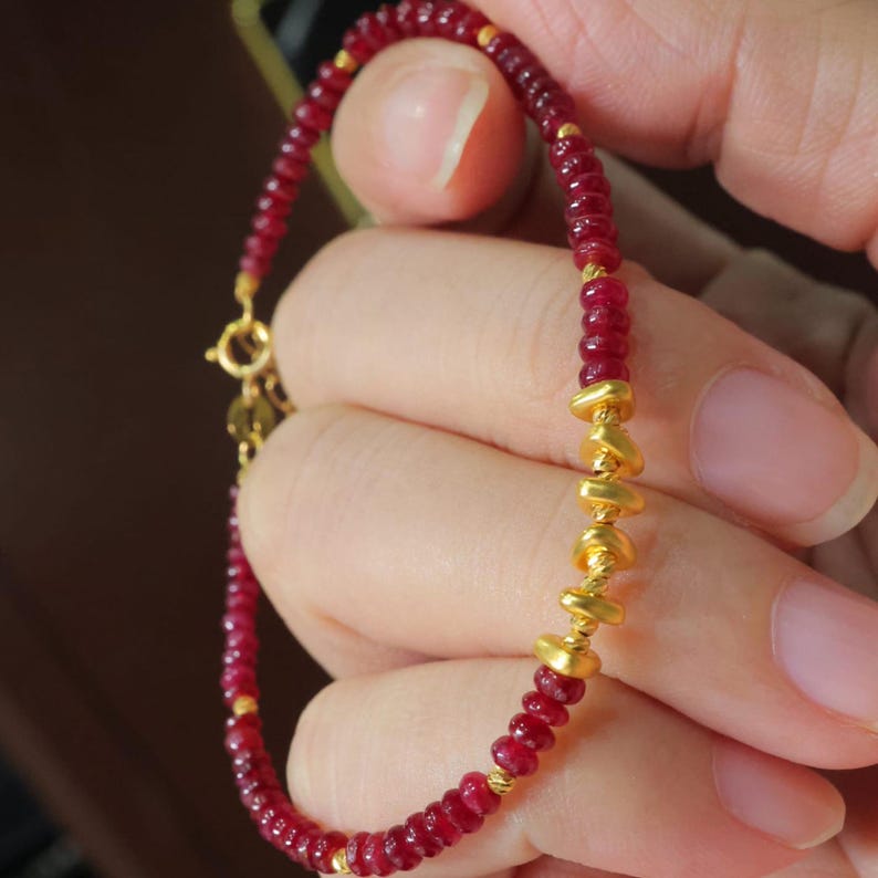 May include: A delicate bracelet featuring small, round, deep red beads interspersed with gold-colored accents. The bracelet has a gold clasp and a section of larger, irregular gold beads. The jewelry is held in a hand.
