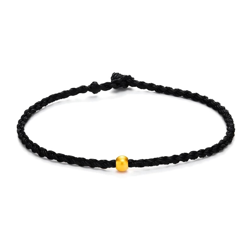 May include: A black braided cord bracelet with a single gold-colored bead in the center. The bracelet is knotted at the ends, and the design is simple and elegant.