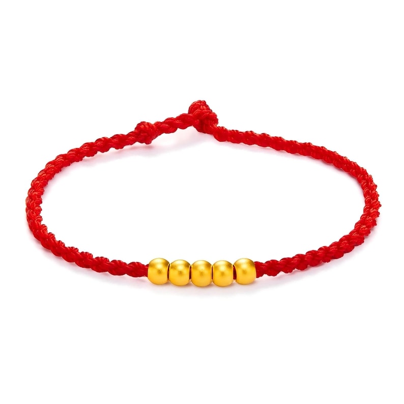 May include: A red braided bracelet with five gold beads. The bracelet is knotted at the ends and is displayed against a white background. The gold beads are evenly spaced along the bracelet.