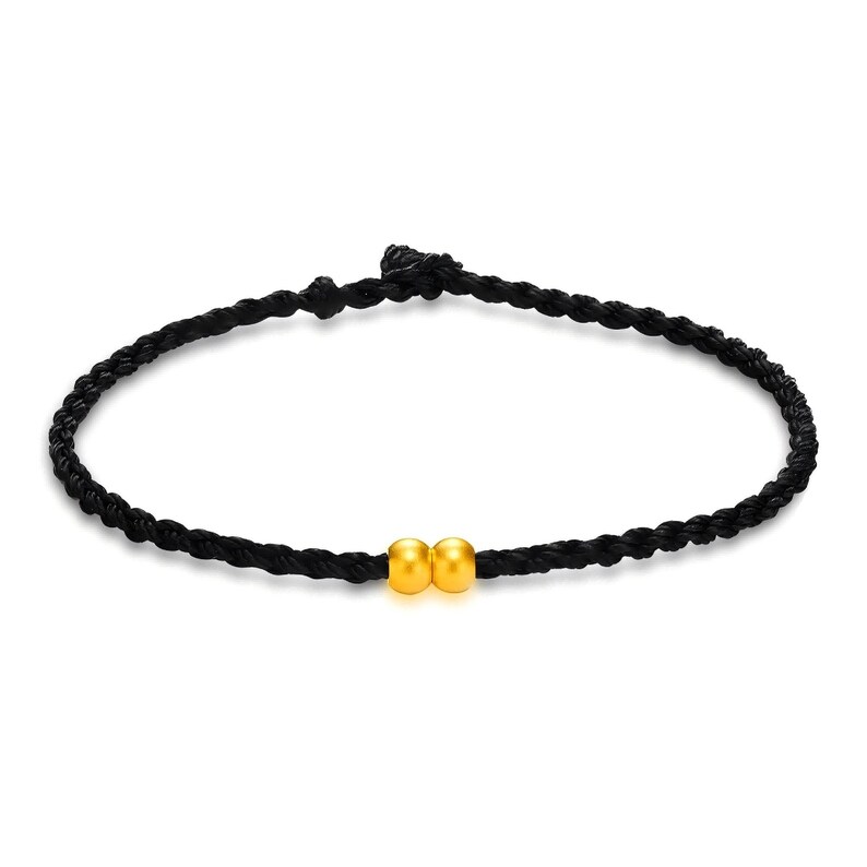 May include: A black braided bracelet featuring two small, round, gold-colored beads. The bracelet is made of a dark, possibly nylon, cord. The beads are centered on the bracelet.
