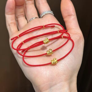 Puede incluir: Tres pulseras de cordón rojo con dijes de cerdo dorados. Cada pulsera presenta un pequeño y detallado dije de cerdo dorado centrado en un cordón rojo vibrante. Las pulseras son sencillas y elegantes.