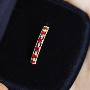 Könnte beinhalten: Ein goldfarbener Ring mit einer Reihe leuchtend roter Edelsteine und kleineren klaren Steinen. Der Ring ist vor einem schwarzen Samthintergrund platziert, der sein Funkeln und Design hervorhebt. Der Ring ist ein Schmuckstück.