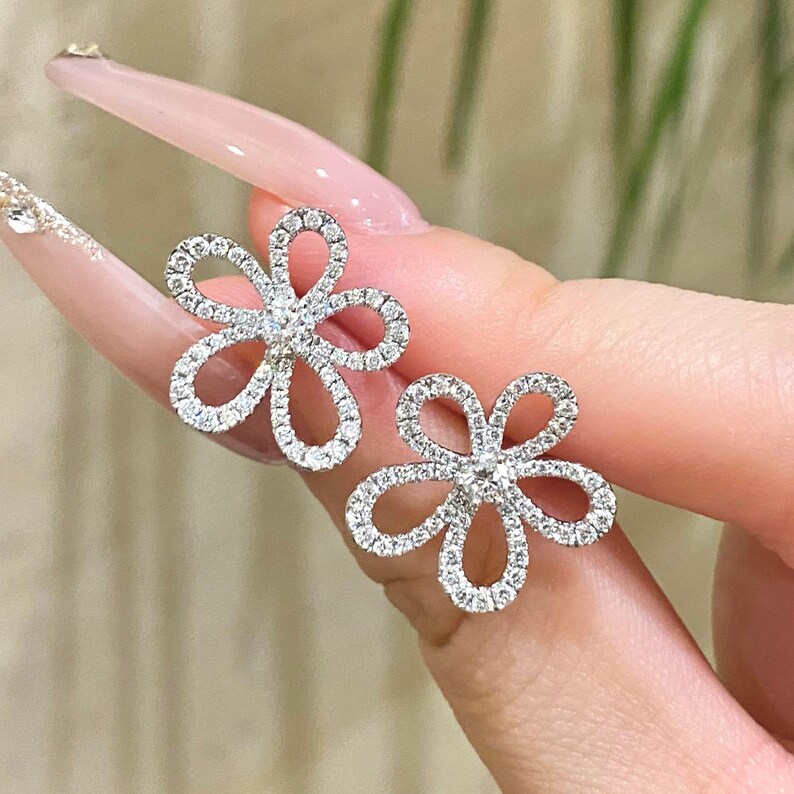 Puede incluir: Un par de pendientes de bot&oacute;n con forma de flor. Cada pendiente presenta un dise&ntilde;o floral con cinco p&eacute;talos, incrustados con peque&ntilde;as piedras brillantes. Los pendientes est&aacute;n engarzados en un metal color plata, creando una apariencia delicada y elegante.
