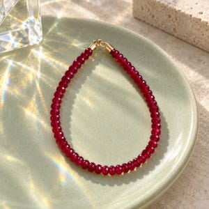 May include: A delicate bracelet composed of small, round, faceted deep red beads. The strand is secured with a polished gold-tone metal clasp at the top, creating a circular shape. The beads exhibit a consistent, vibrant crimson hue throughout.