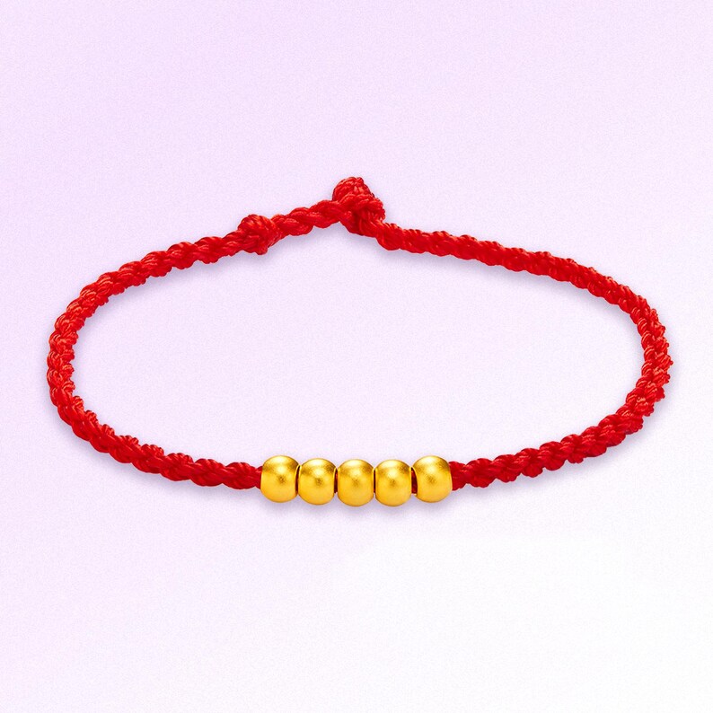 May include: A red braided bracelet with five gold beads. The bracelet is knotted at the ends and is displayed against a soft, light purple background. The gold beads are evenly spaced along the bracelet.