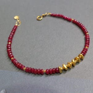 May include: A delicate bracelet featuring small, round, deep red beads interspersed with tiny gold-colored beads. Several larger, flat, gold-colored beads are clustered together. The bracelet has a gold-colored clasp and chain.