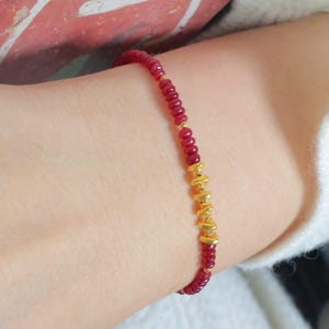 May include: A delicate bracelet featuring small, round, red beads strung together. The bracelet is accented with gold-colored cylindrical beads. The jewelry is worn on a wrist, showcasing the contrast between the red and gold elements.