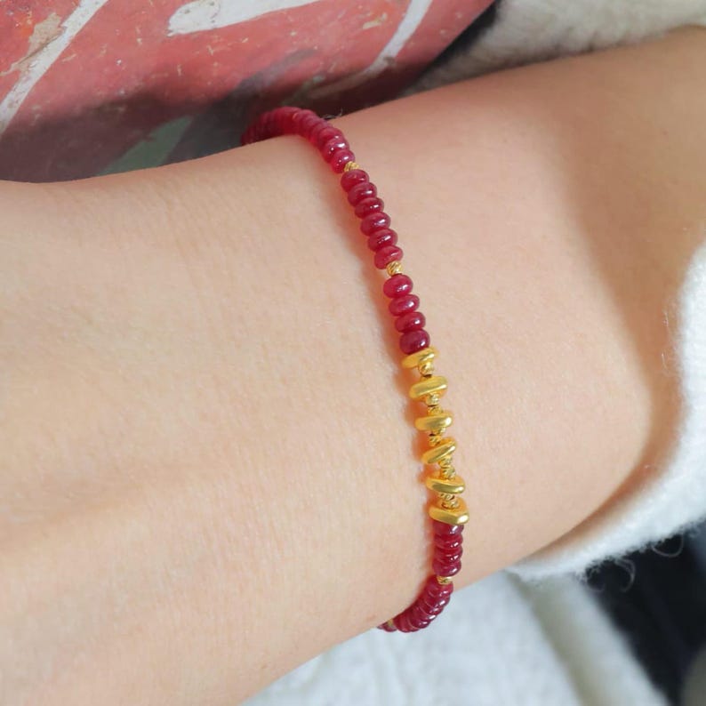 May include: A delicate bracelet featuring small, round, red beads strung together. The bracelet is accented with gold-colored cylindrical beads. The jewelry is worn on a wrist, showcasing the contrast between the red and gold elements.