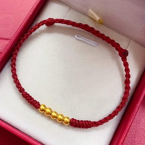 Può includere: Un braccialetto rosso intrecciato con quattro perline dorate. Il braccialetto è presentato all'interno di un portagioie rosso con interno bianco. Il braccialetto è annodato ad entrambe le estremità.