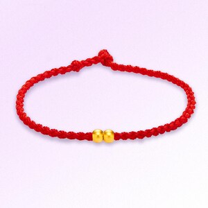 May include: A red braided bracelet with two small, round, gold-colored beads. The bracelet is knotted at the ends and has a simple, elegant design. The background is a soft, gradient purple.