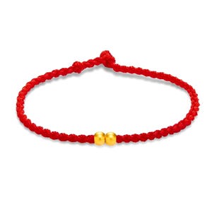 May include: A red braided bracelet with two small, gold-colored beads. The bracelet is knotted at the ends and appears to be made of a soft, flexible material. The beads are round and shiny, adding a touch of elegance to the simple design.