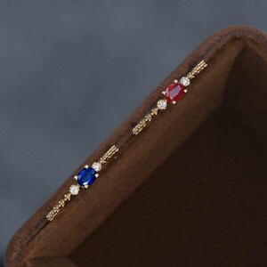 May include: A pair of gold-colored earrings, each featuring an oval-shaped gemstone in either red or blue, accented with small, clear stones. The earrings have a linear design, with the gemstones and smaller stones set along a thin, straight bar.