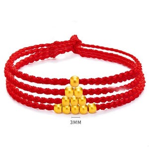 May include: Red braided bracelet with gold-colored beads. The bracelet has three strands of red cord and a pyramid of gold beads in the center. The beads are approximately 3mm in size.