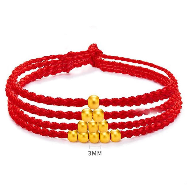May include: Red braided bracelet with gold-colored beads. The bracelet has three strands of red cord and a pyramid of gold beads in the center. The beads are approximately 3mm in size.