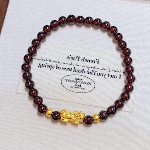 以下が含まれることがあります： 小さな丸い深紅色のビーズで作られたブレスレット。ブレスレットには金色のアクセントと詳細な金のチャームが含まれています。ブレスレットは、テキストが書かれた白い布の上に表示されています。